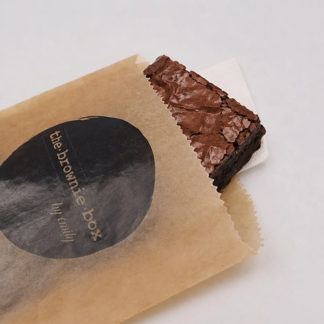 Brown Kraft Paper Counter Bag With Black Circular Branding of The Brown Box Holding A Brownie On A Wooden Countertop Brown Kraft Paper Counter Bag With Black Circular Branding of The Brown Box Holding A Brownie On A Wooden Countertop