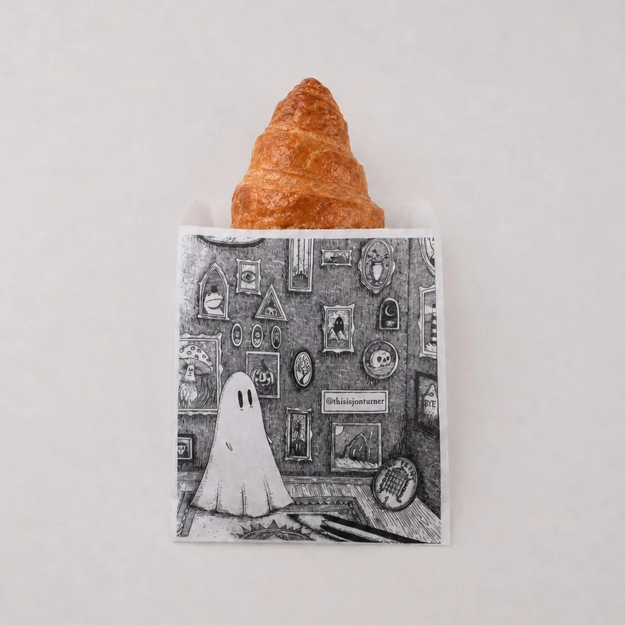 Printed Grey Paper Counter Bag With Ghost Illustration Holding A Croissant On A Light Stone Surface Printed Grey Paper Counter Bag With Ghost Illustration Holding A Croissant On A Light Stone Surface